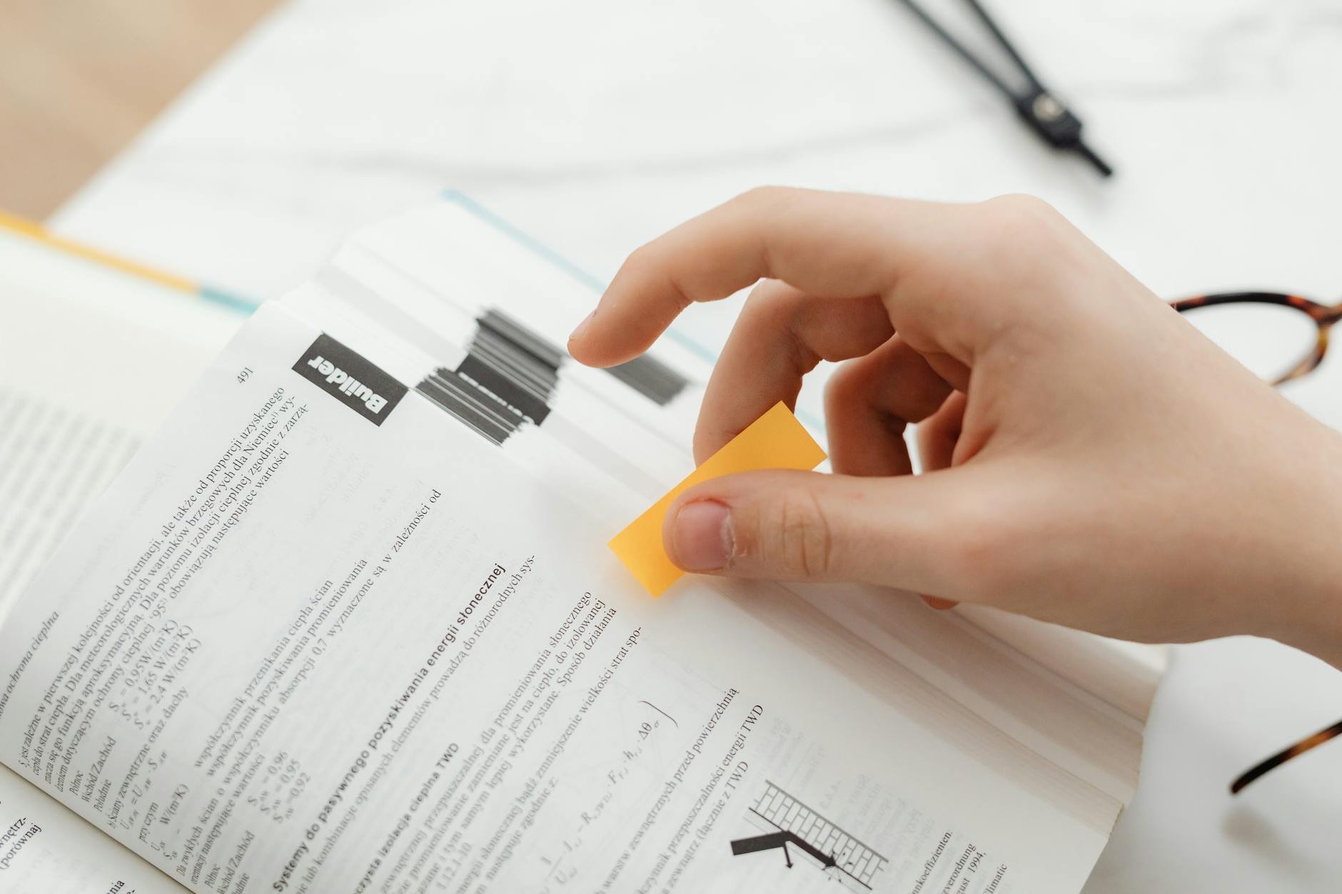 Mão colocando um post-it em livro aberto, simbolizando estudo e otimização.